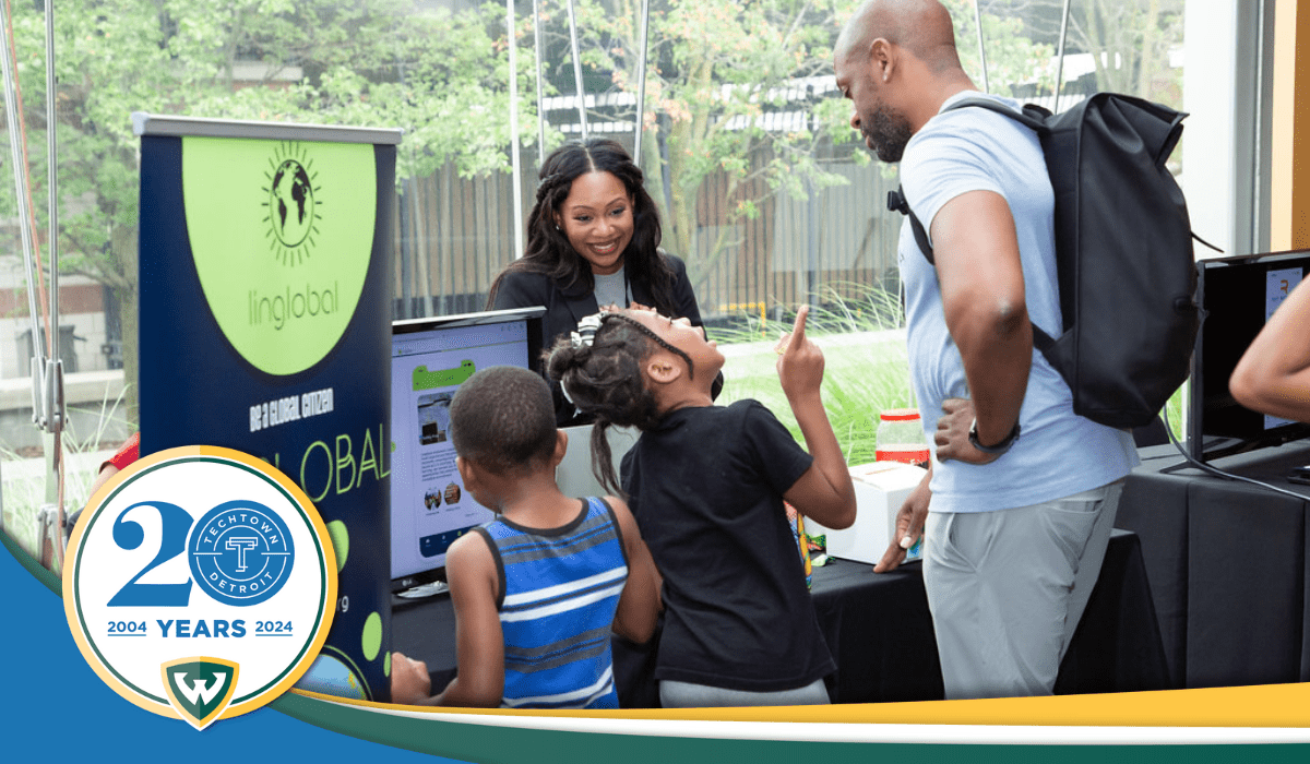 A photo of tech founder Ernestine Lyons, creator of Linglobal, stands behind a table display for her startup. In front of it are two kids and adult looking at the items on the table