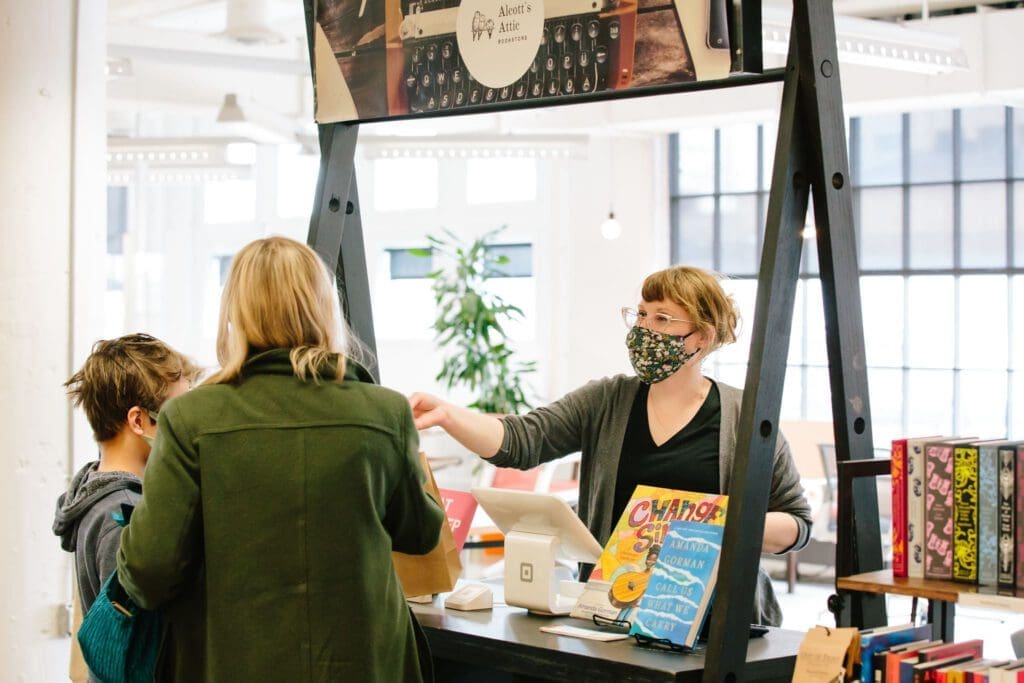 A woman wearing a face masks and standing behind a display fixture of books hands a brown paper bag to a child and woman.