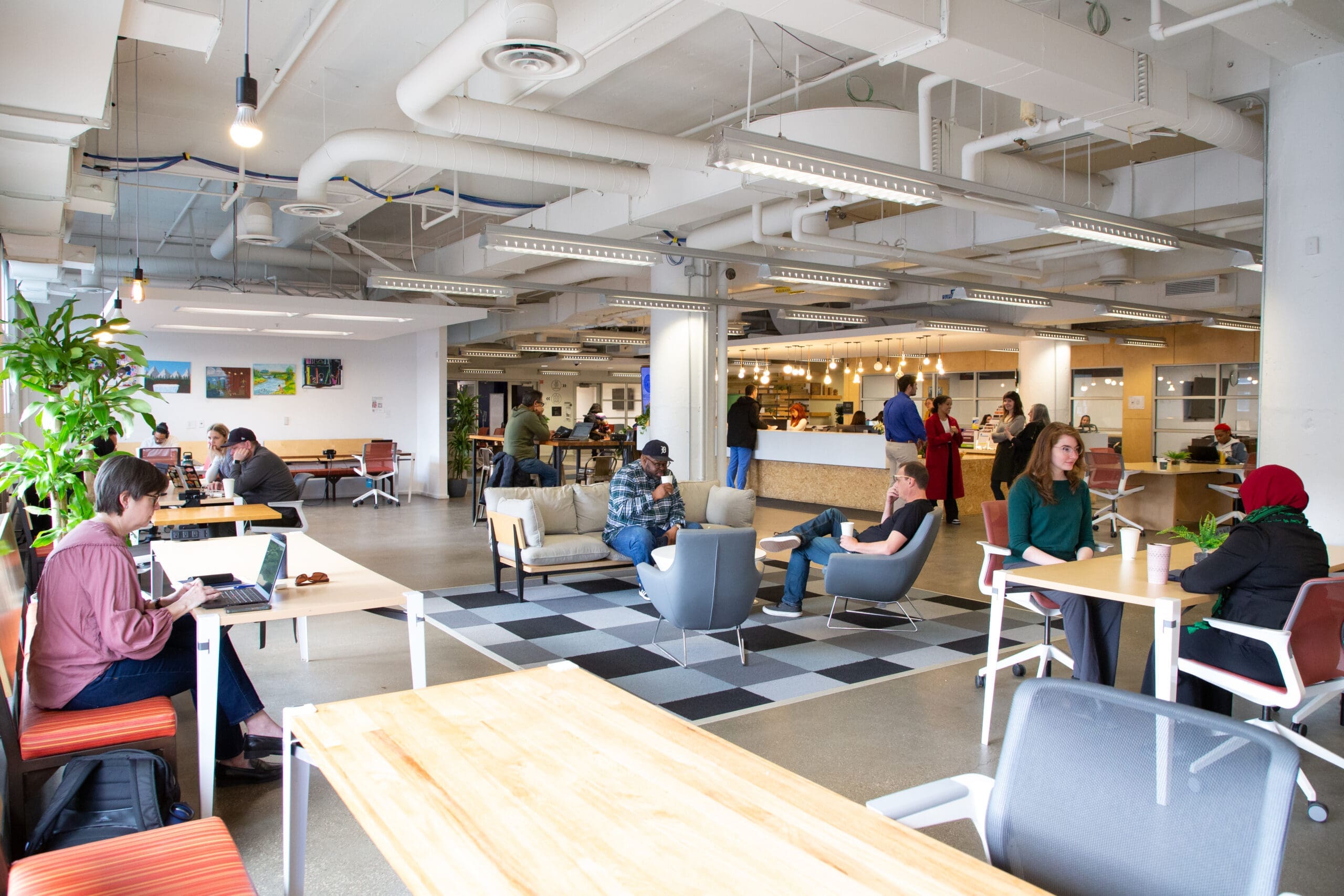 Interior of a spacious office with a modern design, featuring a variety of seating areas including booths and communal tables. Individuals are seen engaging in various activities such as eating, working on laptops and conversing.