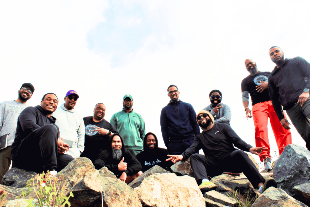 A group of men smile and pose for a photograph. They are standing outside on a rocky area.