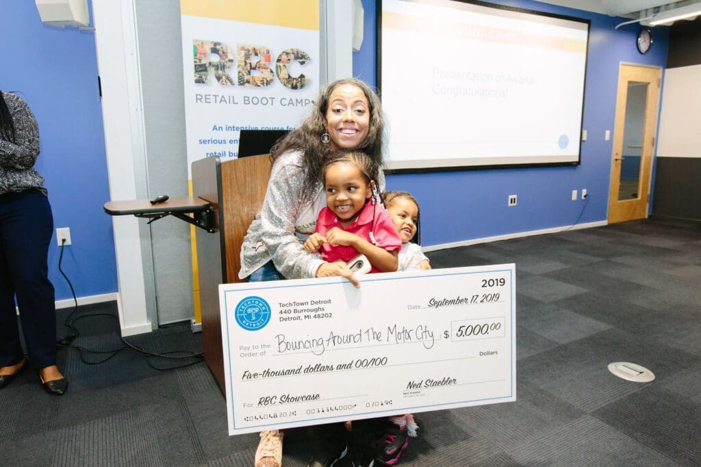 A woman smiles for a camera while holding her two little girls and a $5,000 check for her business Bouncing Around The Motor City
