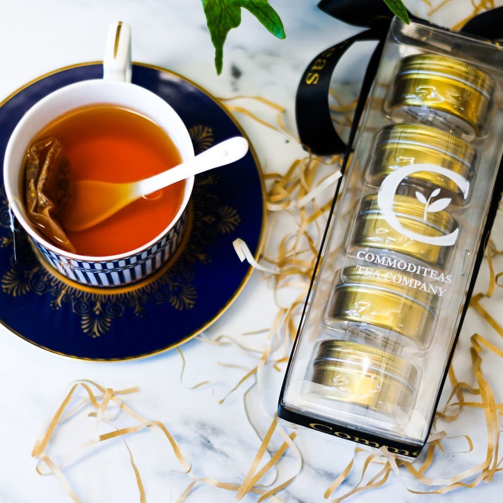 A cup of tea on a saucer with a tea bag in it. Next to the cup is a package of loose-leaf teas from CommodiTeas