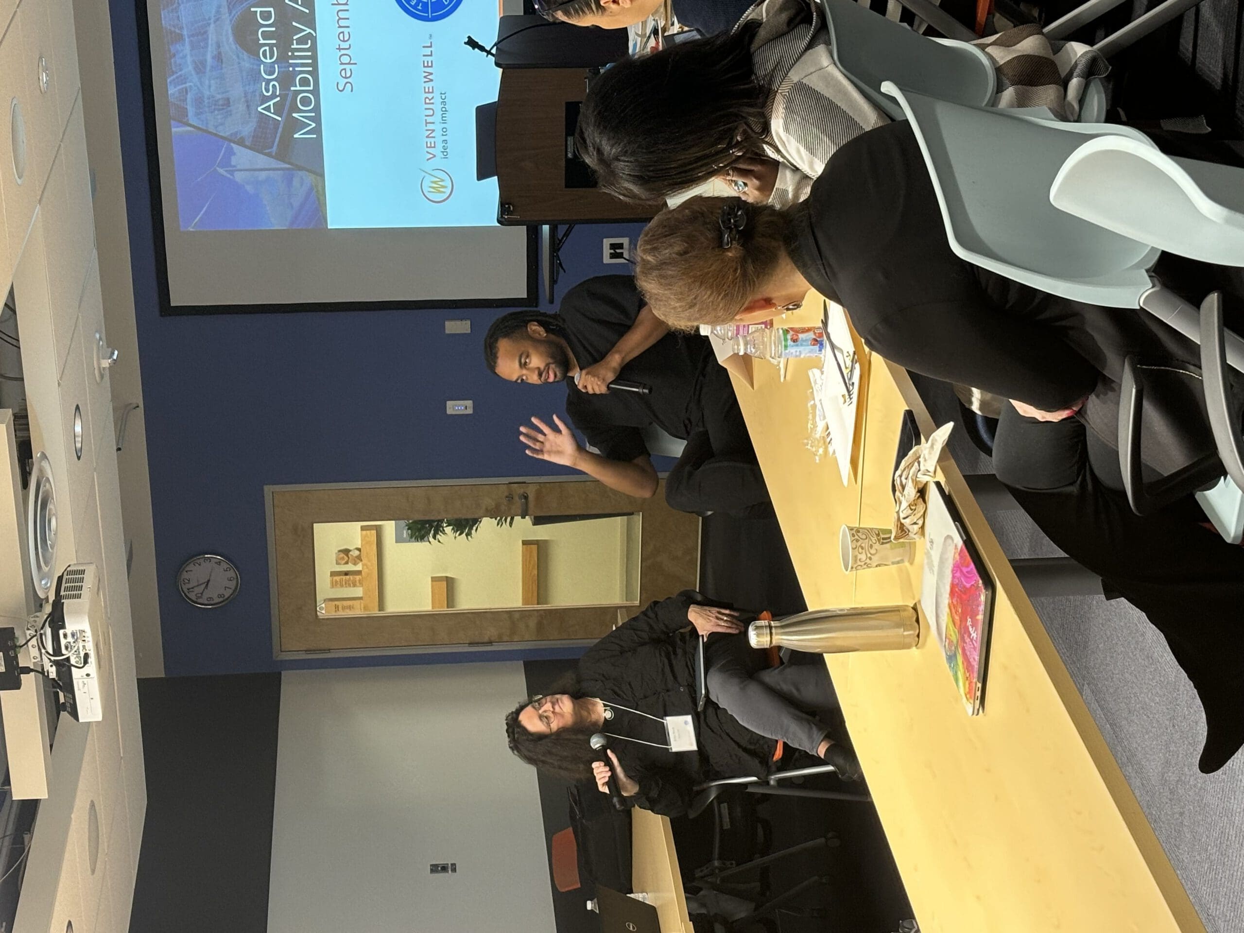 Two people with microphones sit, surrounded by people sitting at desks, round-table style, for a workshop in a conference room