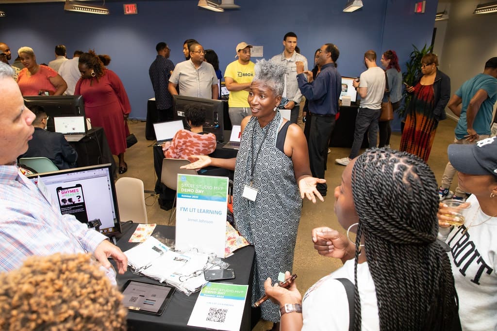 A woman stands behind a table display for her startup I'M LIT Learning. The table has a laptop, tablet and promotional material around it. She is speaking with several people standing around her table.