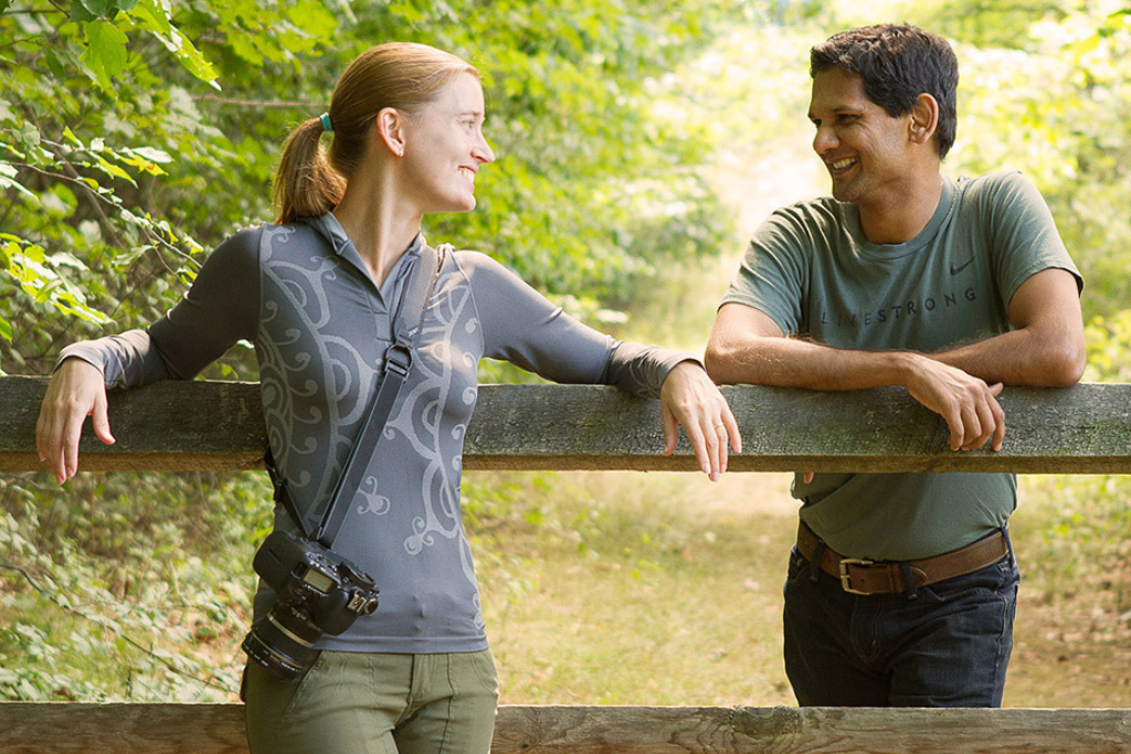 A woman and a man stand outside leaning on a wooden fence. They are smiling at each other. The woman has a camera connected to a camera strap around her shoulder