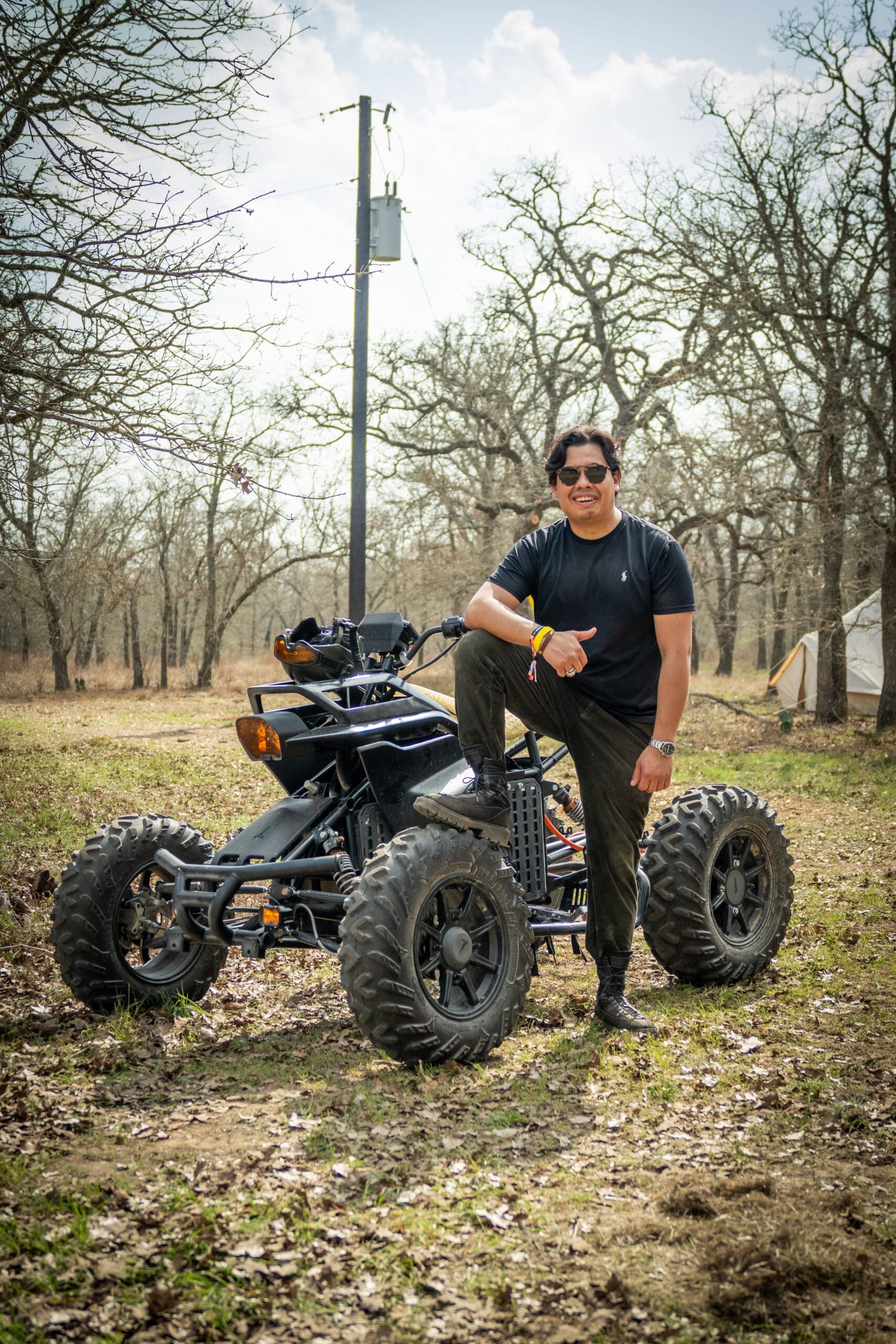 A person smiles while posing beside an all-electric ATV. The person and ATV are outside on a grassy landscape surrounded by trees