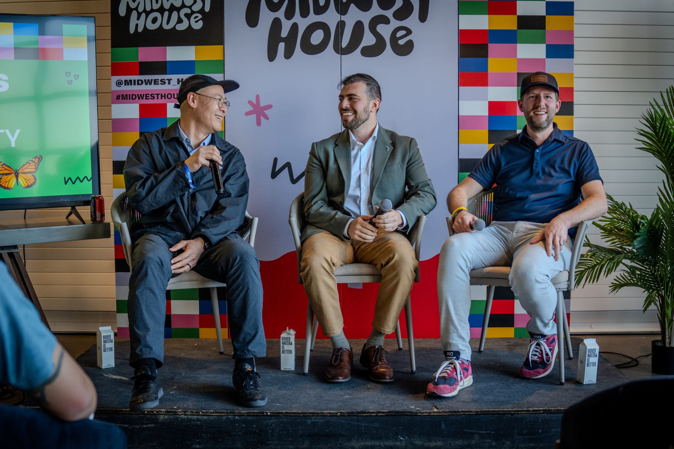 Three people sit in a line of chairs, seated on a stage. Behind them is signage for Midwest House