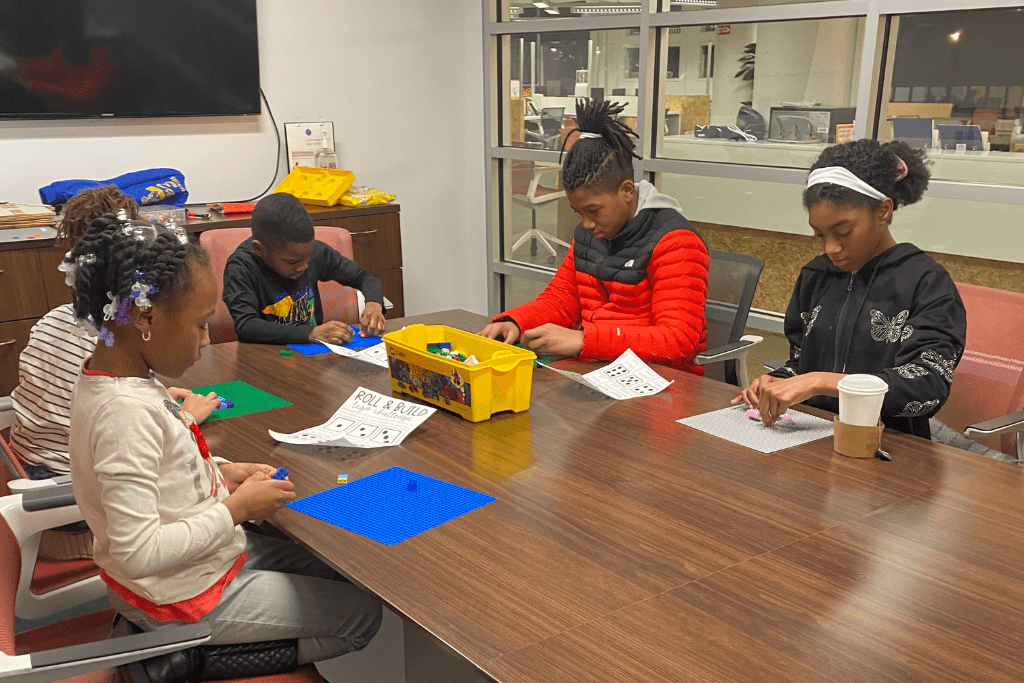 Five children sit around a large table in a conference room, working with learning toys and coloring sheets