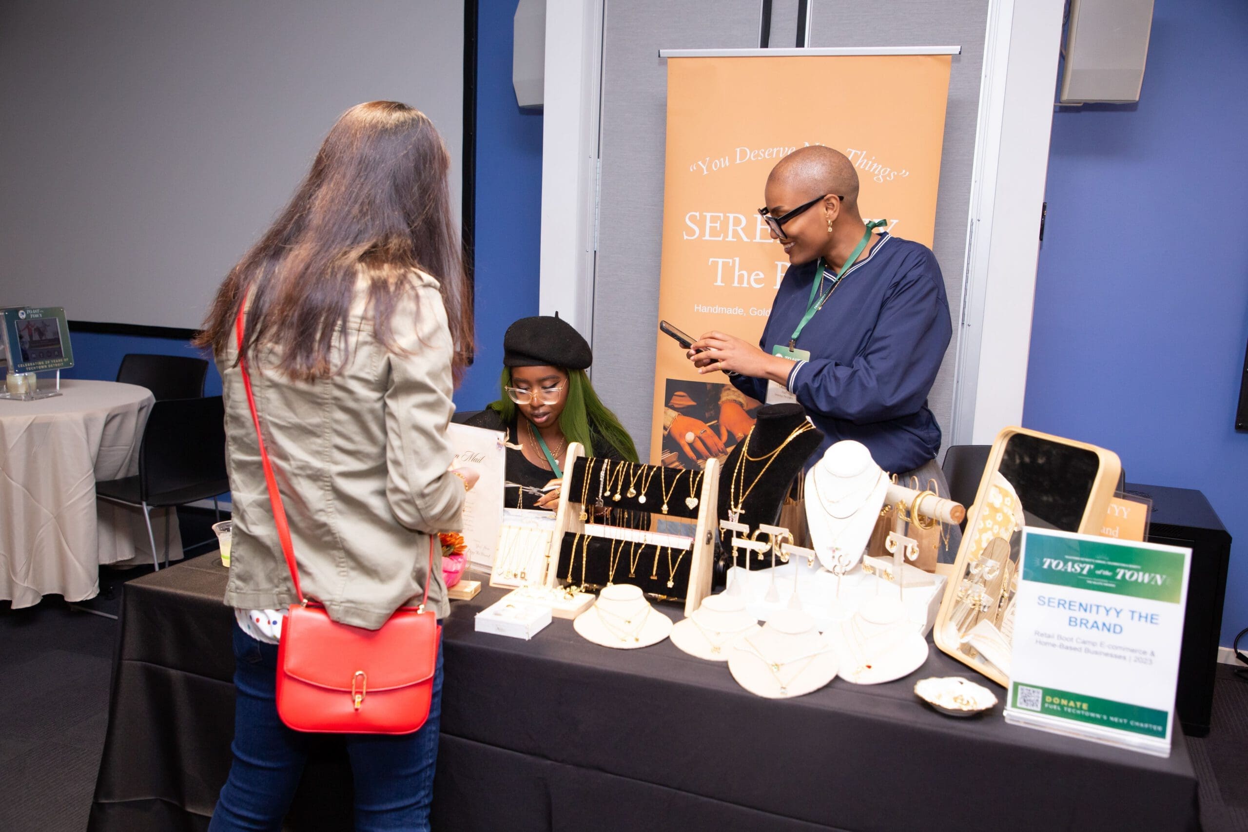 Three people stand around a table display for the jewelry business, Serenityy the Brand. One person is standing in front of the table looking at the products. Another person is sitting down, using pliers to work on a chain. The third person is smiling as she looks at her phone