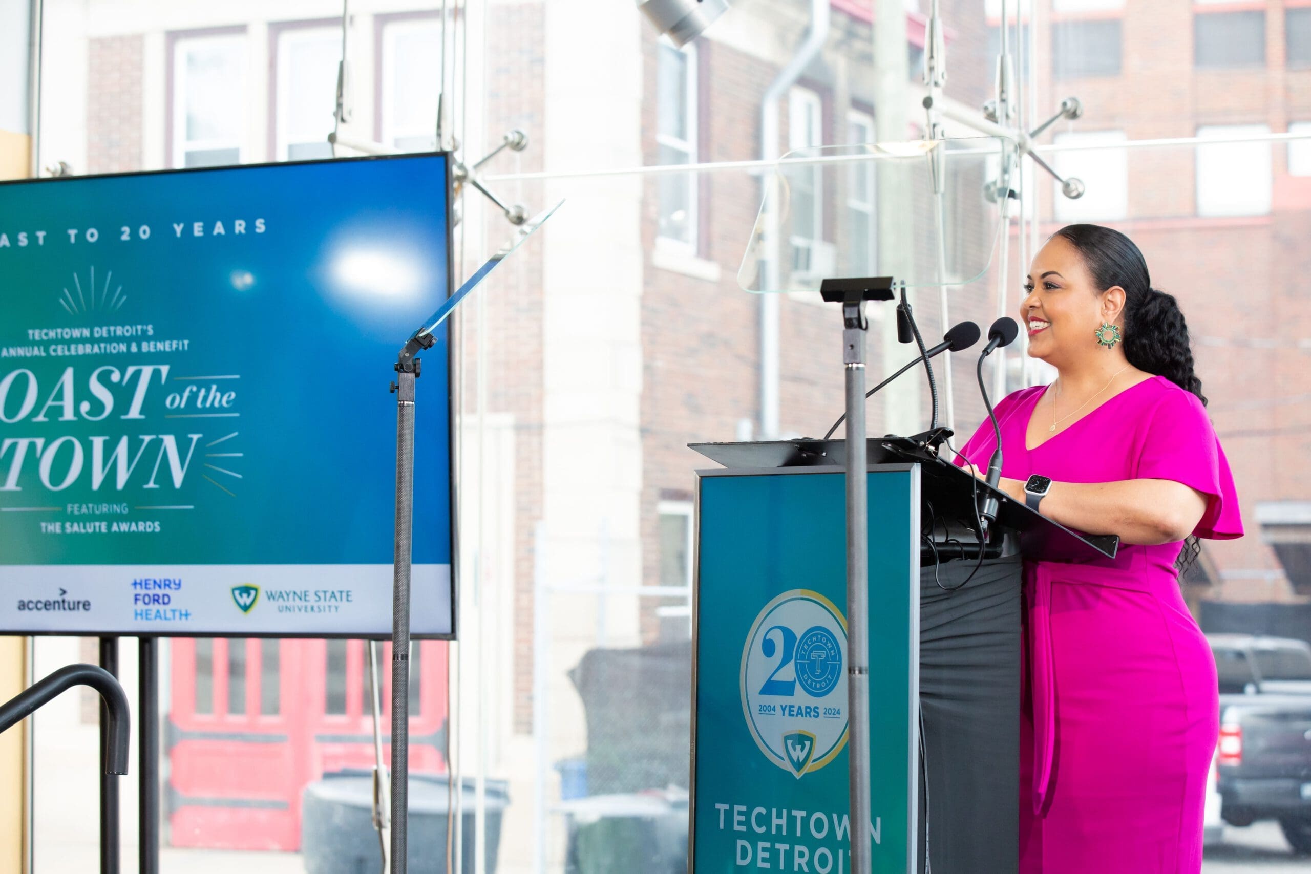 Tati Amare smiles as she stands behind a podium on an atrium stage. Both the podium and large monitor to the right of her feature slides on Toast of the Town and TechTown Detroit